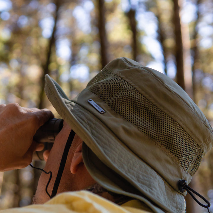 Person wearing a beige hat with a mesh design, holding binoculars in a forest setting.