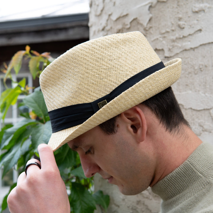 Man wearing a straw hat with a black band, adjusting it outdoors.