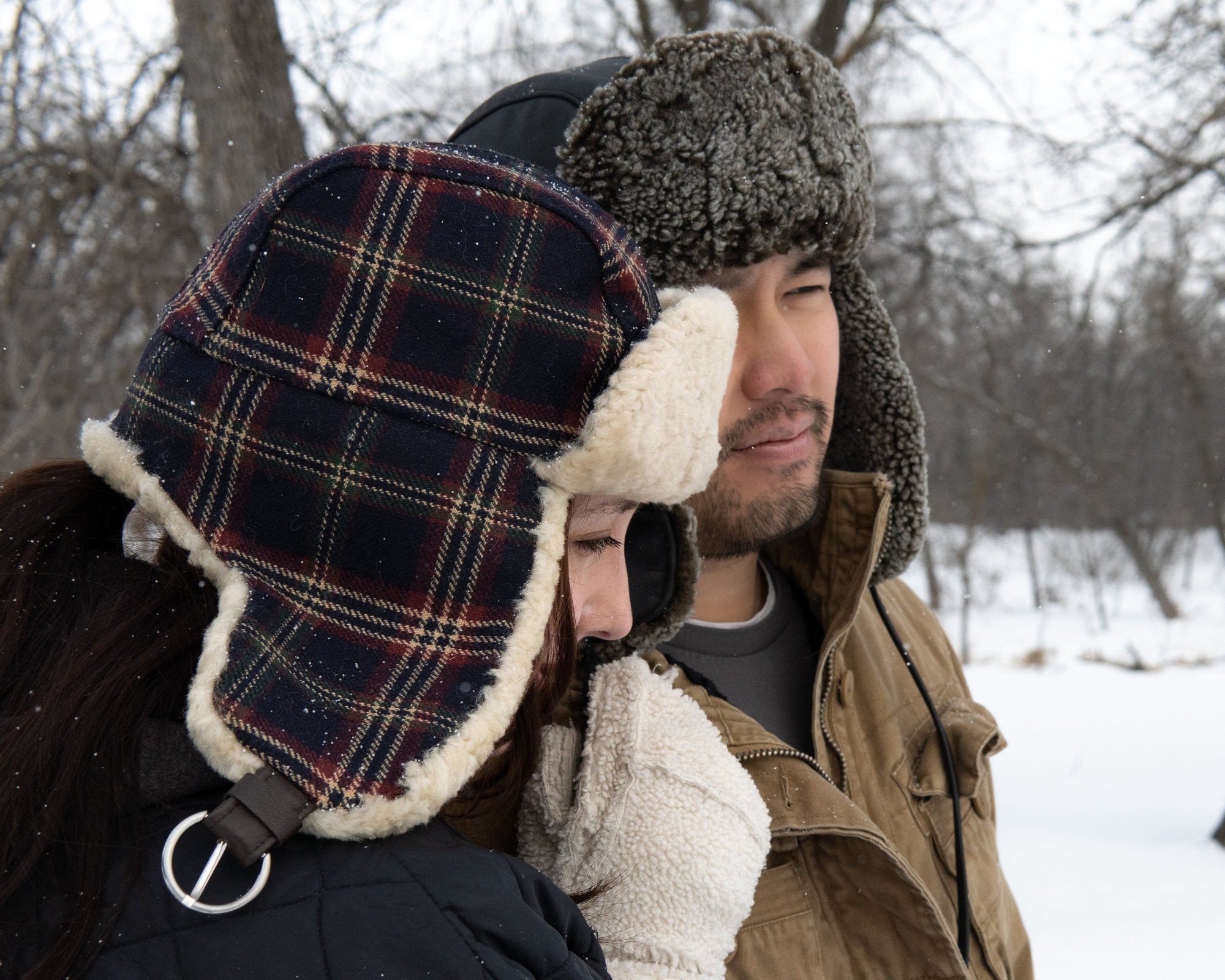 Two people wearing winter hats and coats in a snowy forest.
